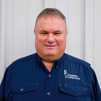 A man smiling in front of a plain, light-colored wall, wearing a dark blue shirt with "Farmers Cooperative" logo, conveying a welcoming and professional vibe.