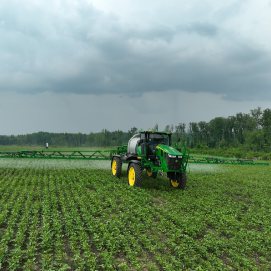 A John Deere tractor spraying a field under a dark, cloud covered sky.