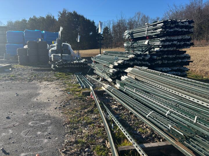 Stacked metal beams and construction materials are outdoors on a sunny day against a backdrop of trees and grass. The setting appears industrial.