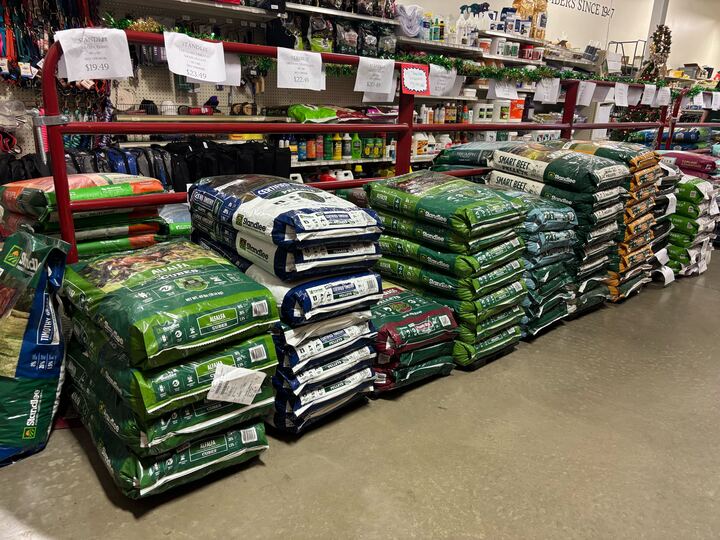 Bags of soil and fertilizer are neatly stacked in rows on a store floor with shelves displaying gardening products above, creating an organized, functional scene.