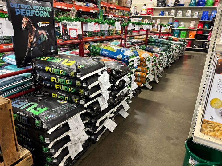 Stacks of horse feed bags are neatly organized in an aisle of a store. A sign featuring a horse promotes performance recovery. Shelves display various feed bags and containers, creating a tidy, well-stocked atmosphere.